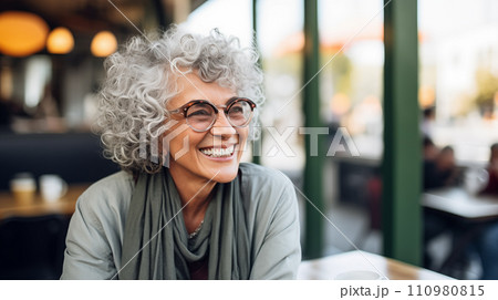 Generative AI The image shows an elderly woman with curly gray hair, wearing glasses and smiling in a cafe setting. 110980815