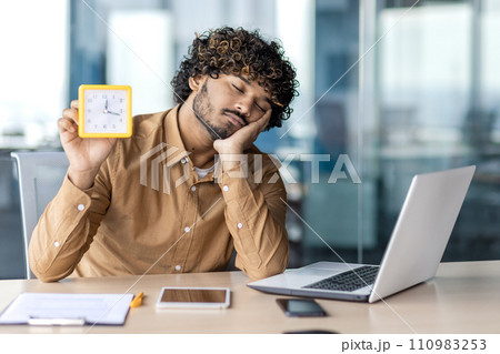 Exhausted muslim man sitting at laptop with closed eyes and showing alarm clock in one hand. Young male in shirt sleeping at office desk during break and holding timer for not be late to meeting. Exhausted muslim man sitting at laptop with closed eyes and showing alarm clock in one hand. Young male in shirt sleeping at office desk during break and holding timer for not be late to meeting. 110983253