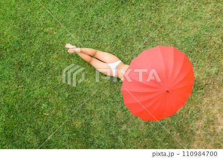 Woman in white bikini lying under red umbrella on the green grass sunbathes at summer day. Top view, drone, aerial view. Woman in white bikini lying under red umbrella on the green grass sunbathes at summer day. Top view, drone, aerial view. 110984700