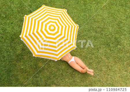 Woman in white bikini lying under yellow umbrella on the green grass sunbathes at summer day. Top view, drone, aerial view. Woman in white bikini lying under yellow umbrella on the green grass sunbathes at summer day. Top view, drone, aerial view. 110984725