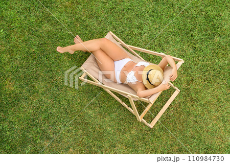 Woman in a white bikini in hat with laptop sitting on deck chair on the green grass sunbathes at summer day. Top view, drone, aerial view. 110984730