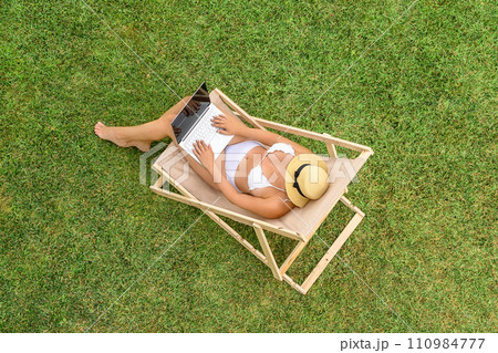 Woman in a white bikini sitting on deck chair with laptop on the green grass sunbathes at summer day. Top view, drone, aerial view. Woman in a white bikini sitting on deck chair with laptop on the green grass sunbathes at summer day. Top view, drone, aerial view. 110984777