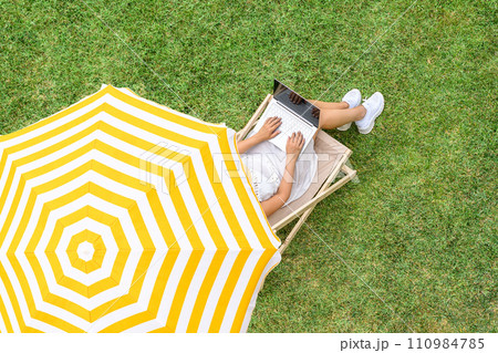 Woman in a white dress sitting on deck chair under yellow umbrella with laptop on the green grass sunbathes at summer day. Top view, drone, aerial view. Woman in a white dress sitting on deck chair under yellow umbrella with laptop on the green grass sunbathes at summer day. Top view, drone, aerial view. 110984785