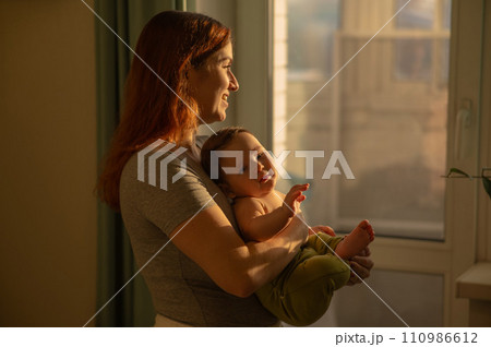 Caucasian woman tenderly holds her newborn son while standing near the window. Caucasian woman tenderly holds her newborn son while standing near the window. 110986612