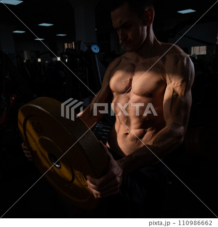 Muscular shirtless man doing exercise with weight plate in gym. Muscular shirtless man doing exercise with weight plate in gym. 110986662
