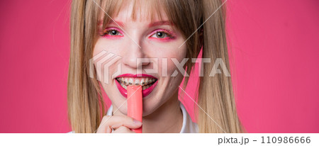 Portrait of a young woman with braces and bright makeup chewing gum on a pink background. 110986666