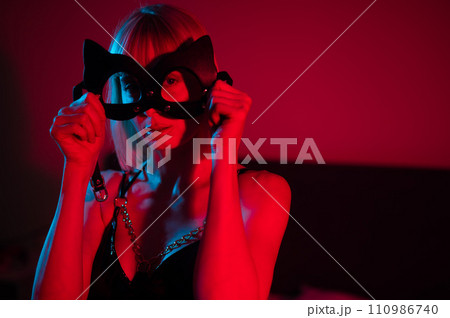 Blonde woman putting on leather mask in bedroom in red blue neon light. Blonde woman putting on leather mask in bedroom in red blue neon light. 110986740