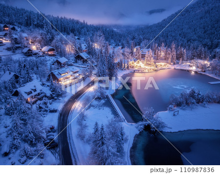 Aerial view of snowy village, trees, lake, street lights at night Aerial view of snowy village, trees, lake, street lights at night 110987836
