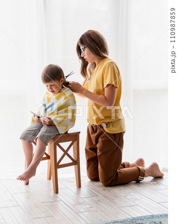 Mother cuts her son's hair at home. Little boy sits covered with cloth and holds pair of scissors. Self care at home. Candid lifestyle. 110987859