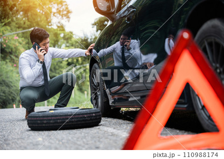 Broken down car with a red triangle warning sign on the road. Businessman waiting for assistance while leaning on the car wheel. Concept of car breakdown and roadside rescue service. 110988174