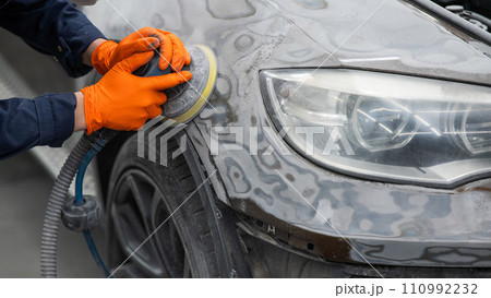 A mechanic sands the putty on a car body with a machine. Repair after an accident. A mechanic sands the putty on a car body with a machine. Repair after an accident. 110992232