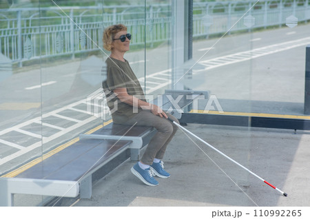 An elderly blind woman is waiting for transport at a bus stop. 110992265