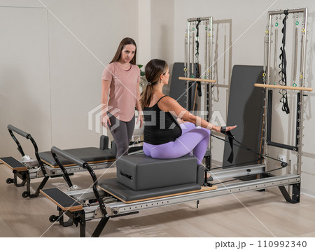 Caucasian pregnant woman doing Pilates exercises on a reformer machine with an individual trainer.  110992340