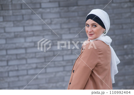 Portrait of a young blue-eyed woman in a hijab against a gray brick wall. A Muslim woman looks at the camera turning around. 110992404