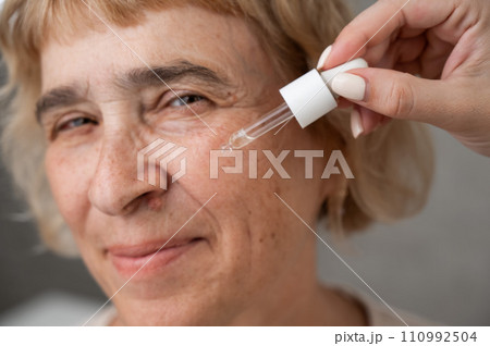 Close-up portrait of an old woman applying hyaluronic acid serum with a pipette. Anti-aging face care. Close-up portrait of an old woman applying hyaluronic acid serum with a pipette. Anti-aging face care. 110992504
