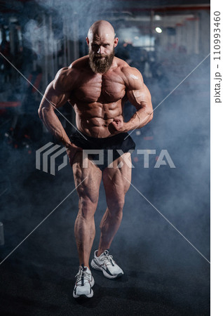 Muscular bald man posing in shorts. Bodybuilder showing off his shape in the gym. Muscular bald man posing in shorts. Bodybuilder showing off his shape in the gym. 110993460