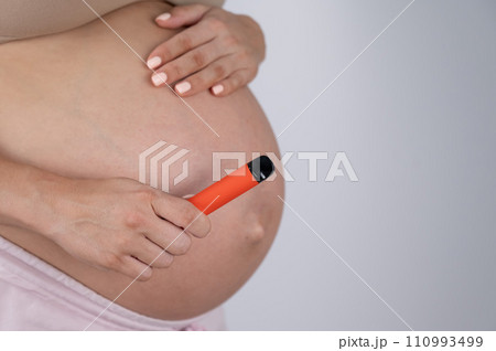 A pregnant woman smokes a vape. A girl holds an electronic cigarette against the background of her bare tummy. Copy space. A pregnant woman smokes a vape. A girl holds an electronic cigarette against the background of her bare tummy. Copy space. 110993499