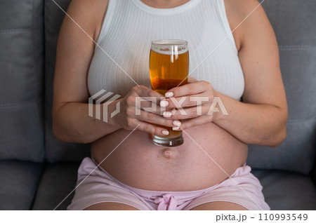 Close-up of the belly of a pregnant woman holding a glass of beer while sitting on the sofa. Close-up of the belly of a pregnant woman holding a glass of beer while sitting on the sofa. 110993549
