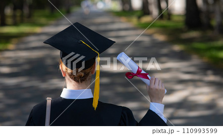 Rear view of a caucasian woman in a graduate gown holding a diploma outdoors.  110993599