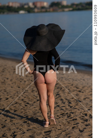 Rear view of woman in large straw hat and black swimsuit posing on the beach. Rear view of woman in large straw hat and black swimsuit posing on the beach. 110993609