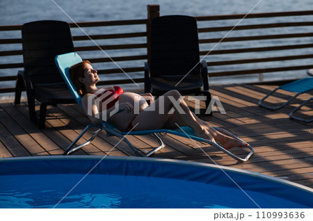 Red-haired pregnant woman in a red bikini sunbathes lying on a sun lounger next to the pool. Red-haired pregnant woman in a red bikini sunbathes lying on a sun lounger next to the pool. 110993636