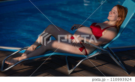 Red-haired pregnant woman in a red bikini sunbathes lying on a sun lounger next to the pool. 110993924
