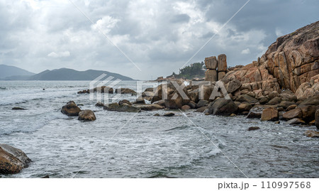 Coastal waves crash against monumental granite boulders lining shore. majestic seascape features vast expanse of rocks, under turbulent, cloud-laden sky, untamed natural panorama 110997568