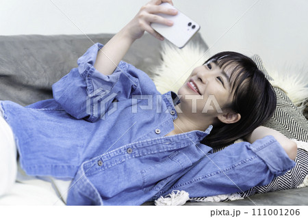 ソファに寝転んでスマホ動画を観る女性 111001206