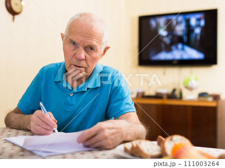 Elderly man writes a letter on a sheet of paper while sitting at a table in room 111003264