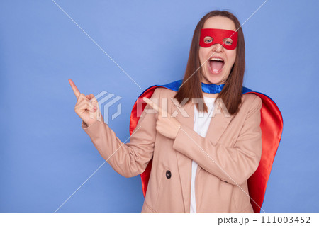 Amazed happy brown haired woman wearing superhero costume posing isolated over blue background exclaiming and pointing both fingers away at advertising area 111003452