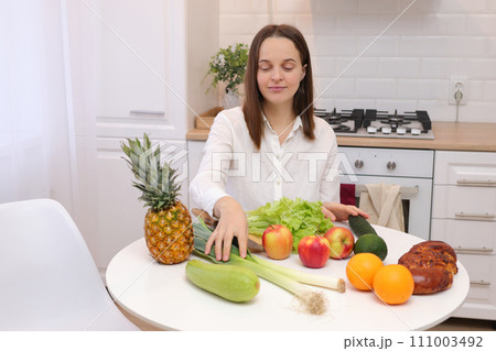 Healthy lifestyle. Raw vegan. Domestic kitchen. Nutritious dinner. Caucasian brown haired woman sitting at kitchen table with fruit and vegetables cooking organic meals at home Healthy lifestyle. Raw vegan. Domestic kitchen. Nutritious dinner. Caucasian brown haired woman sitting at kitchen table with fruit and vegetables cooking organic meals at home 111003492