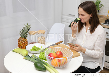 Confused puzzled Caucasian brown haired woman sitting at kitchen table with fruit and vegetables smelling not fresh cucumber from refrigerator or freezer 111003502