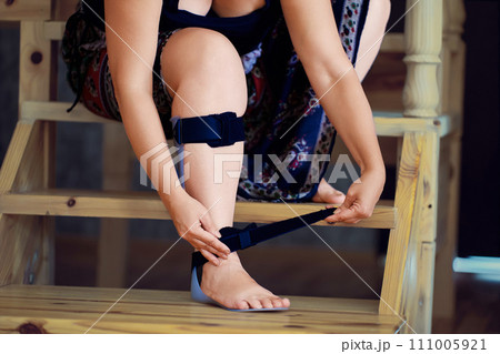 Woman in skirt puts on solid ankle foot orthosis on her injured leg, sitting on wooden step of an interfloor staircase at home. Woman in skirt puts on solid ankle foot orthosis on her injured leg, sitting on wooden step of an interfloor staircase at home. 111005921