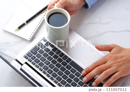 机にノートパソコンやノートを広げ、コーヒーを飲みながらデスクワークをしている男性の両手のイメージ 111007012
