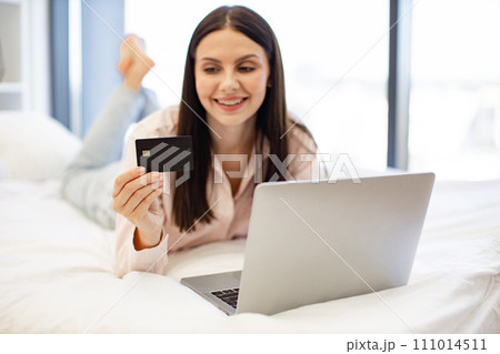 Charming woman in casual attire enjoying online shopping while lying on bed. Charming woman in casual attire enjoying online shopping while lying on bed. 111014511
