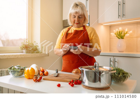 Woman housewife holds phone using cooking apps websites search recipes, chatting with friend during food preparation. Female cooking vegetables in cozy wooden kitchen n Woman housewife holds phone using cooking apps websites search recipes, chatting with friend during food preparation. Female cooking vegetables in cozy wooden kitchen n 111014580