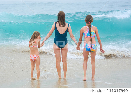 Mother and children playing on the ocean beach. Family enjoying the ocean. Mother holds girls's hands and they all look at the ocean together. 111017940