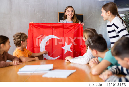 Preteens pupils looking at national flag of Turkey while learning different cultures with positive young female teacher in schoolroom Preteens pupils looking at national flag of Turkey while learning different cultures with positive young female teacher in schoolroom 111018178