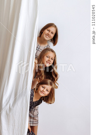 Three girls peek out from behind a curtain against a white wall Three girls peek out from behind a curtain against a white wall 111020545