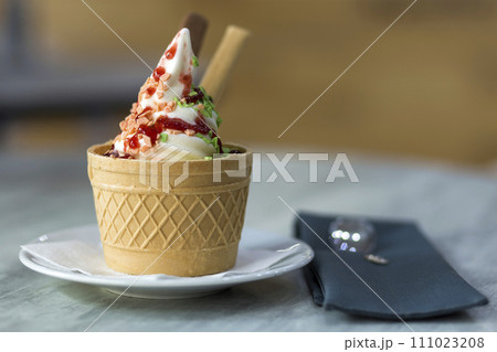 White plate with ice cream dessert in wafer cup with chocolate cookies and creative decoration topping on blurred colorful interior background. 111023208