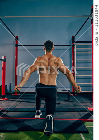 Back training. Athletic, sportive young man training shirtless in gym, doing back exercises with resistance band Back training. Athletic, sportive young man training shirtless in gym, doing back exercises with resistance band 111031844