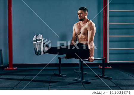 Full-body workout. Muscular, shirtless athletic man training in modern gym, doing L-sit exercise on parallel bars. Full-body workout. Muscular, shirtless athletic man training in modern gym, doing L-sit exercise on parallel bars. 111031871