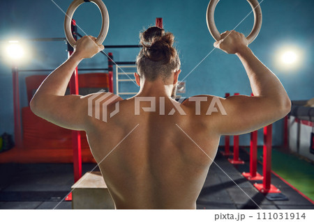 Shirtless, athletic young man showing his muscular, strong body, back, training with gymnastic rings in gym. 111031914