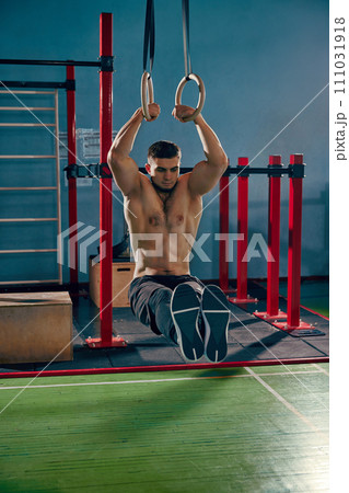 Advanced training. Muscular, athletic young man with relief, strong shirtless body doing L-sit exercises on gymnastic rings in gym Advanced training. Muscular, athletic young man with relief, strong shirtless body doing L-sit exercises on gymnastic rings in gym 111031918