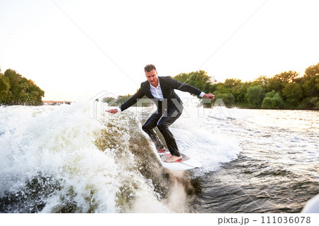 Young man in classic suit rides a wakeboard on the river or lake near city. A careless clerk escaped from a stuffy office to take up his favorite active sport. Best summer leisure after routine work. Young man in classic suit rides a wakeboard on the river or lake near city. A careless clerk escaped from a stuffy office to take up his favorite active sport. Best summer leisure after routine work. 111036078