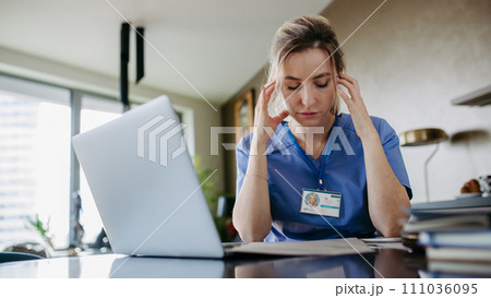 Nurse paying bills online, sitting in living room, working on notebook. Doctor in scrubs dealing with work issues from home, after workday, telemedicine consultation. Work-life balance for healthcare Nurse paying bills online, sitting in living room, working on notebook. Doctor in scrubs dealing with work issues from home, after workday, telemedicine consultation. Work-life balance for healthcare 111036095