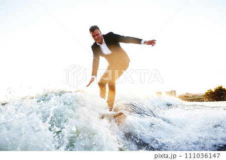 Young man in classic suit rides a wakeboard on the river or lake near city. Clerk escaped from a stuffy office to take up his favorite active sport. Best summer leisure after routine work. Sun flare. 111036147