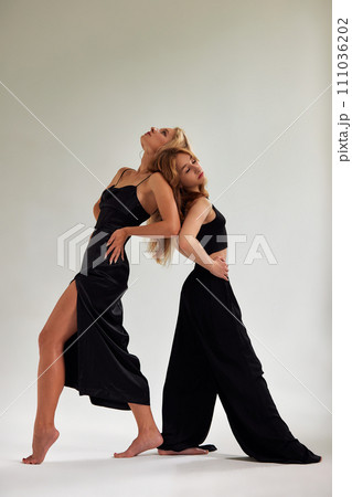 Two young sexy barefoot women dressed in black clothes posing on white background in studio. Stylish blonde girls, one in black dress and the other in black top and loose pants, standing back to back. 111036202