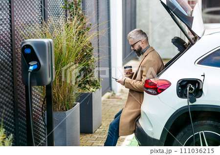 Portrait of businessman waiting while electric car is charging, sitting in car trunk, scrolling on his smartphone, and drinking coffee. An electric vehicle charging station. Portrait of businessman waiting while electric car is charging, sitting in car trunk, scrolling on his smartphone, and drinking coffee. An electric vehicle charging station. 111036215
