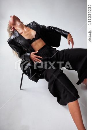 Young sexy woman dressed in black leather jacket, black bra and black loose pants posing on white background in studio. Stylish blonde girl in black clothes sitting on chair leaning back. Top view. 111036239